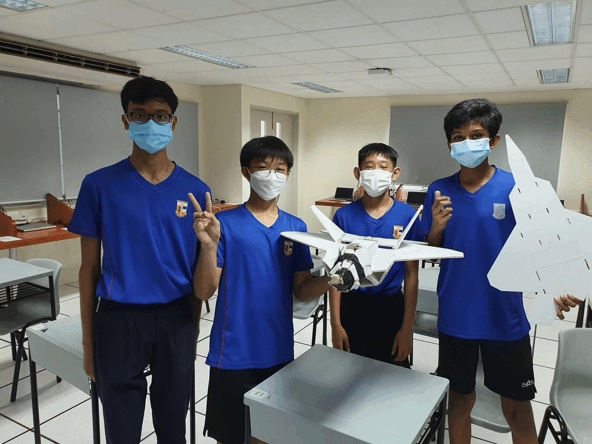 Four masked students in blue uniforms stand in a classroom holding a white model aircraft.